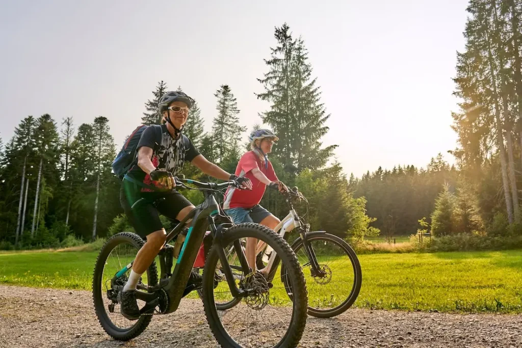 Zwei Frauen sitzen auf einem Mountainbike und fahren auf einem geschottertem Weg. Im Hintergrund ist eine Wiese und ein Wald zu sehen