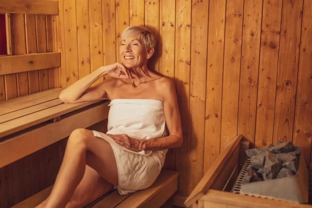 Eine Dame sitzt mit einem weißen Handtuch freundlich schauen in einer Sauna